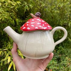 a clay teapot with a red mushroom on top is held in front of some trees