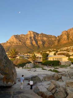 two people are walking on the beach near some rocks and boulders with mountains in the background