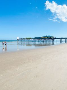 two people are walking on the beach with a pier in the background and blue sky