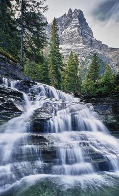 a waterfall in the mountains with trees around it