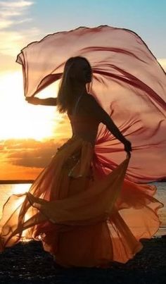 a woman is standing on the beach with her dress blowing in the wind as the sun sets