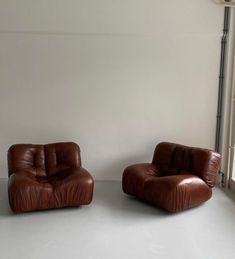 two brown leather chairs sitting next to each other in front of a window on a white floor