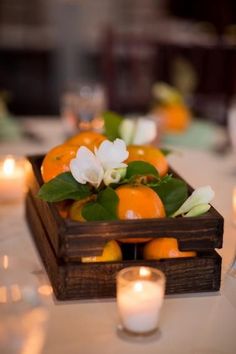 an arrangement of oranges and flowers in a wooden box on a table with candles