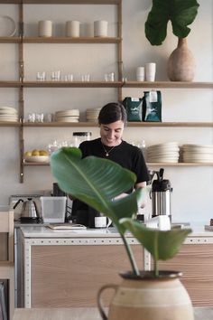 a woman standing in front of a counter filled with coffee cups and saucers next to a plant