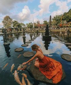 a woman in an orange dress kneeling down to catch fish from the water with her hands