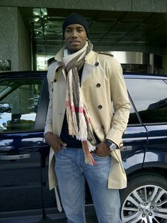 a man standing in front of a car wearing a trench coat, scarf and jeans