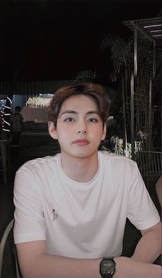 a young man sitting at a table with a cell phone in his hand and wearing a white t - shirt