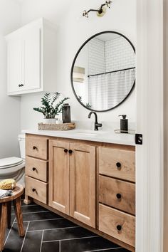 a bathroom with white walls and black tile flooring, wooden cabinets and a round mirror on the wall