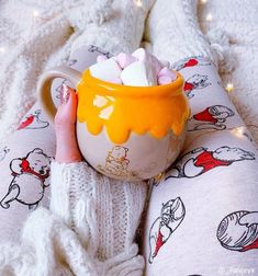 a person is holding a mug with marshmallows in it on the bed