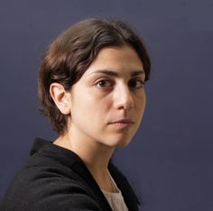 a woman with dark hair and black jacket looking at the camera while standing in front of a blue background