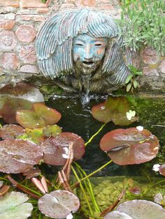 a fountain with water lilies and lily pads in front of a brick wall that has a statue of a woman's head above it