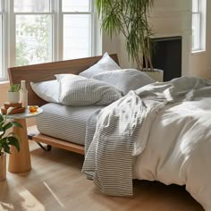 a bed with white sheets and pillows next to a potted plant in front of a window