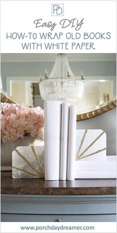 two white books sitting on top of a table next to pink flowers and a mirror