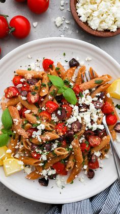 a white plate topped with pasta, tomatoes and feta cheese next to a bowl of olives