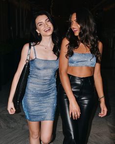 two young women walking down a sidewalk at night, one wearing a blue top and the other black leather pants