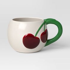 a white cup with two cherries painted on the side and green handles, sitting in front of a gray background