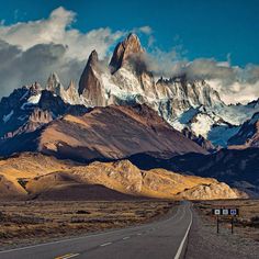 an empty road in the middle of mountains with signs on it and snow capped peaks