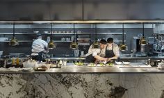 two chefs in a kitchen preparing food on the counter and people working behind them at work