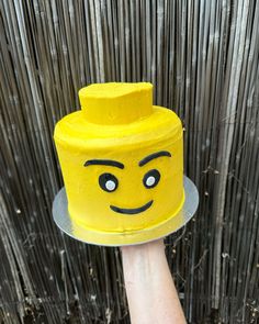 a hand holding a yellow lego head cake on top of a metal platter in front of a bamboo fence