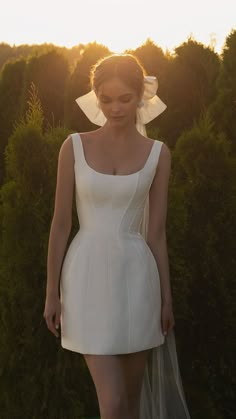 a woman wearing a white dress and veil standing in front of bushes with the sun behind her