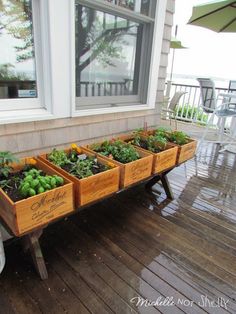 a wooden table topped with lots of plants on top of a porch next to a window