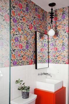 a bathroom with floral wallpaper and orange cabinet under a mirror above a white sink