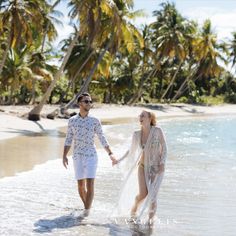 a man and woman walking in the water on a beach with palm trees behind them