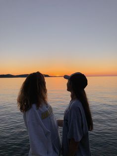 two people standing on the edge of a boat looking out at the water as the sun sets