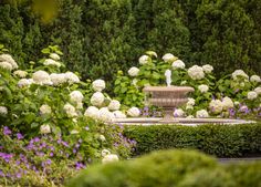 a fountain surrounded by bushes and flowers in the middle of a garden with purple and white flowers