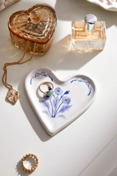 a heart shaped dish sitting on top of a table next to two rings and perfume bottles