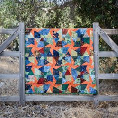 an orange and blue quilt is hanging on a wooden fence in front of some trees