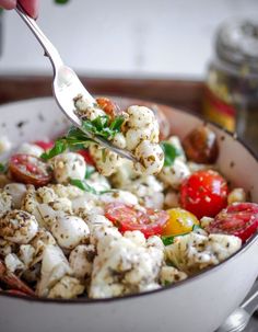 a person holding a spoon over a bowl full of food that includes tomatoes and cauliflower