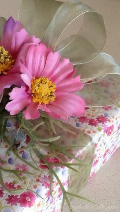 two pink flowers are in a vase on a floral print table cloth with silver ribbon