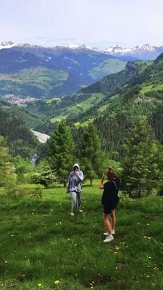 two people standing in the grass with mountains in the background