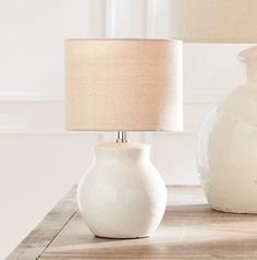 two white vases sitting next to each other on a table with a lamp in front of them