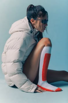 a woman sitting on the ground with her legs crossed wearing white and orange socks, an oversize puffy jacket and black sunglasses