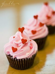 three cupcakes with pink frosting and red hearts on them sitting on a table