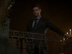 a man in a suit and tie stands on a balcony railing with candles lit behind him