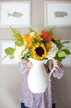 a woman holding a white vase with flowers in it and two pictures on the wall behind her