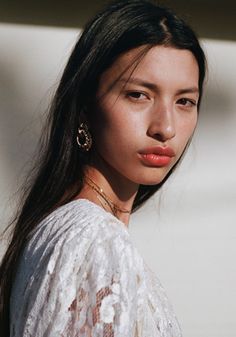 a woman with long black hair wearing gold hoop earrings and a white top is looking off to the side