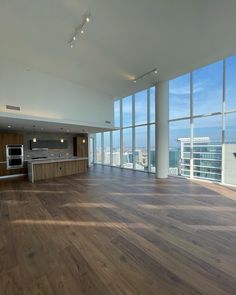 an empty living room with large windows and wood flooring in front of the kitchen