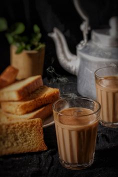 two glasses filled with liquid next to slices of bread and a teapot on a table