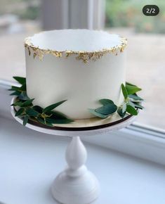 a white cake with gold trim and greenery on top sitting in front of a window