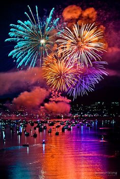 fireworks are lit up in the night sky over water and buildings with lights on them