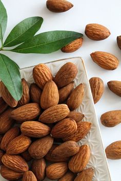 almonds and leaves on a white surface next to a square plate with nuts in it