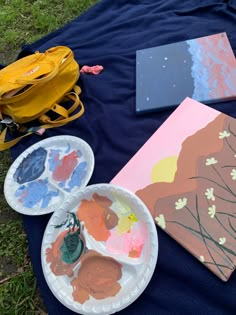 two paper plates with paint on them next to an art book and yellow backpack sitting on the ground