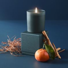 a candle sitting on top of a box next to an orange and some sticks with leaves