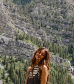 a woman standing in front of a mountain with trees on the side and mountains behind her