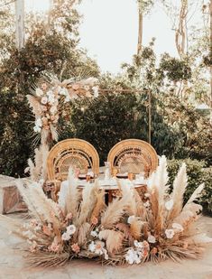 an outdoor table set up with wicker chairs and floral centerpieces in front of greenery