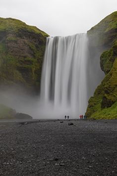 three people standing at the base of a waterfall with green moss growing on the rocks